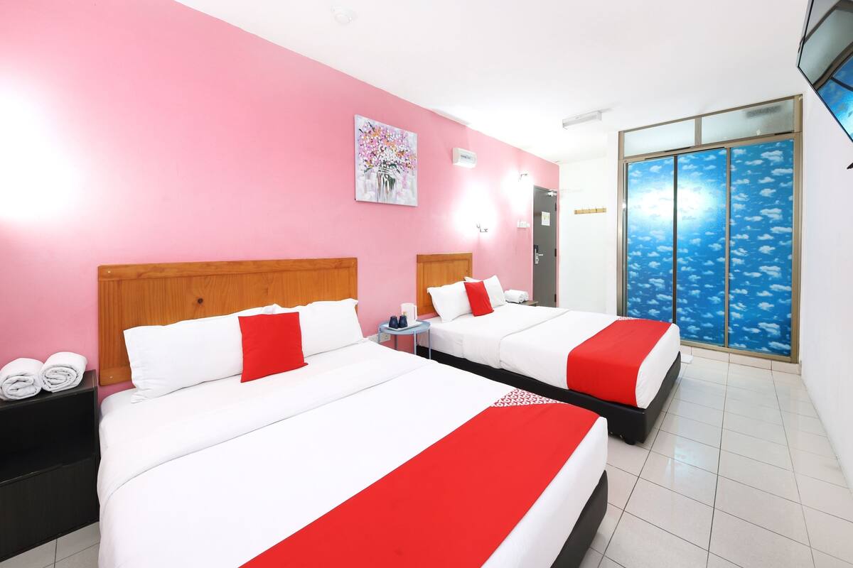 A hotel room with two beds, each with white bedding and red accents, pink walls, a painting, and a large window with a view of the sky.
