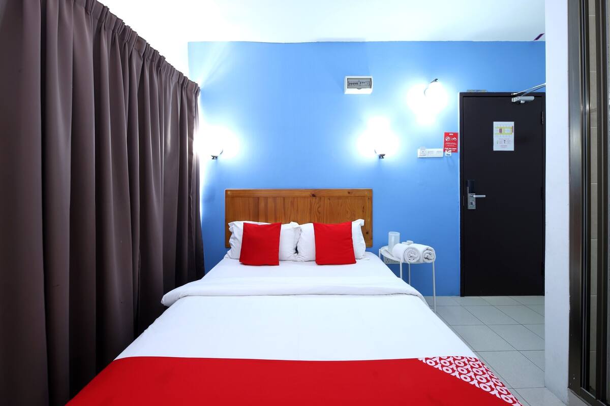 A hotel room with a bed, curtains, a wooden headboard, two wall-mounted lights, a door, and a wall-mounted thermostat.