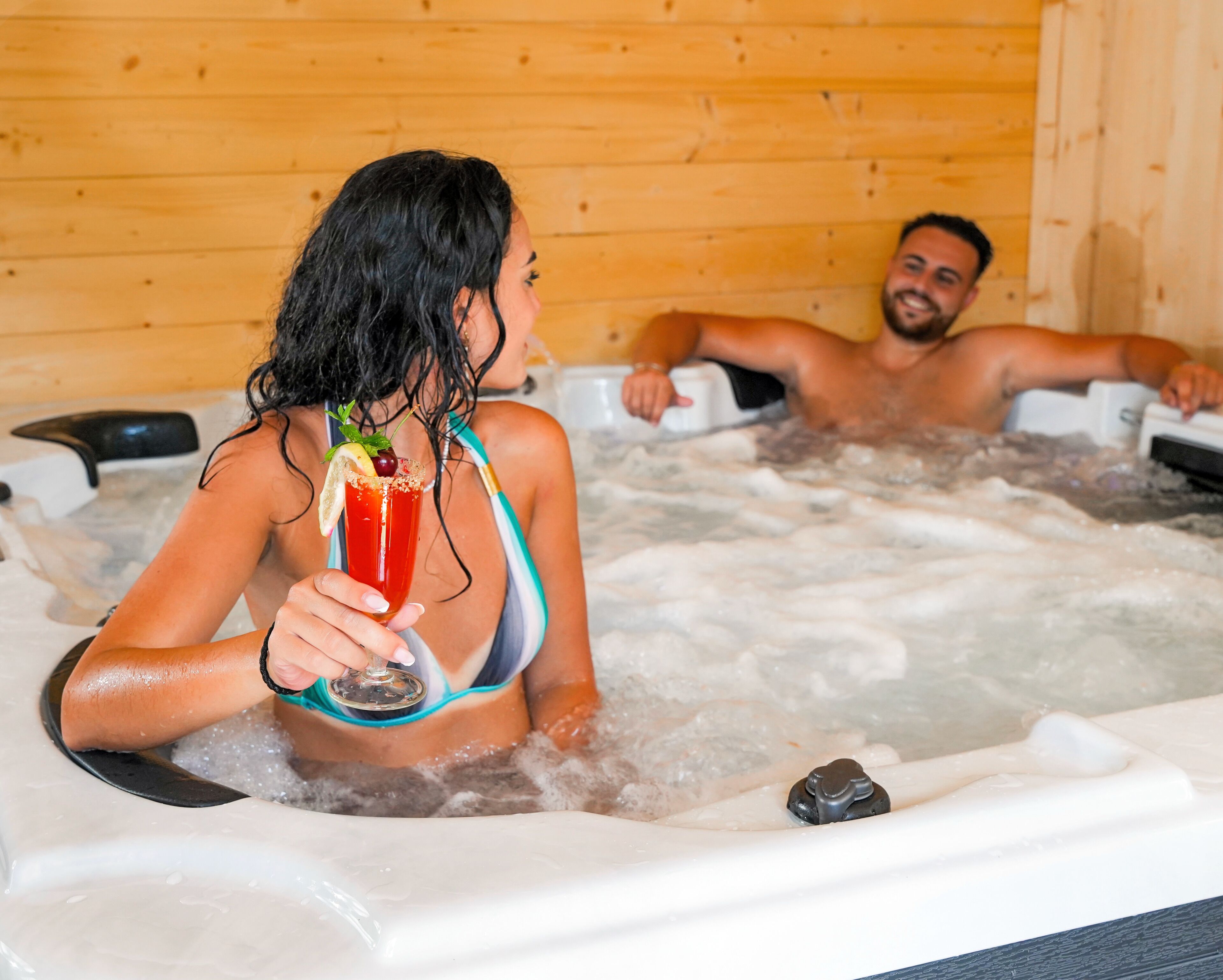 Two people in a hot tub with wooden walls, holding drinks.