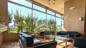 A modern lounge area with leather seating, a glass coffee table, and large windows offering a view of trees and buildings.