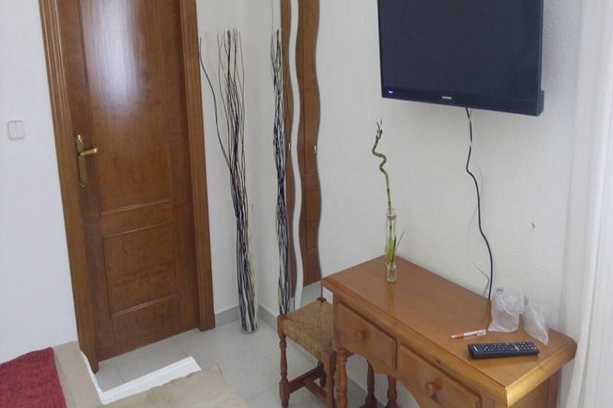 A room with a wooden door, a television mounted on the wall, a wooden desk with a vase of flowers, and a chair.