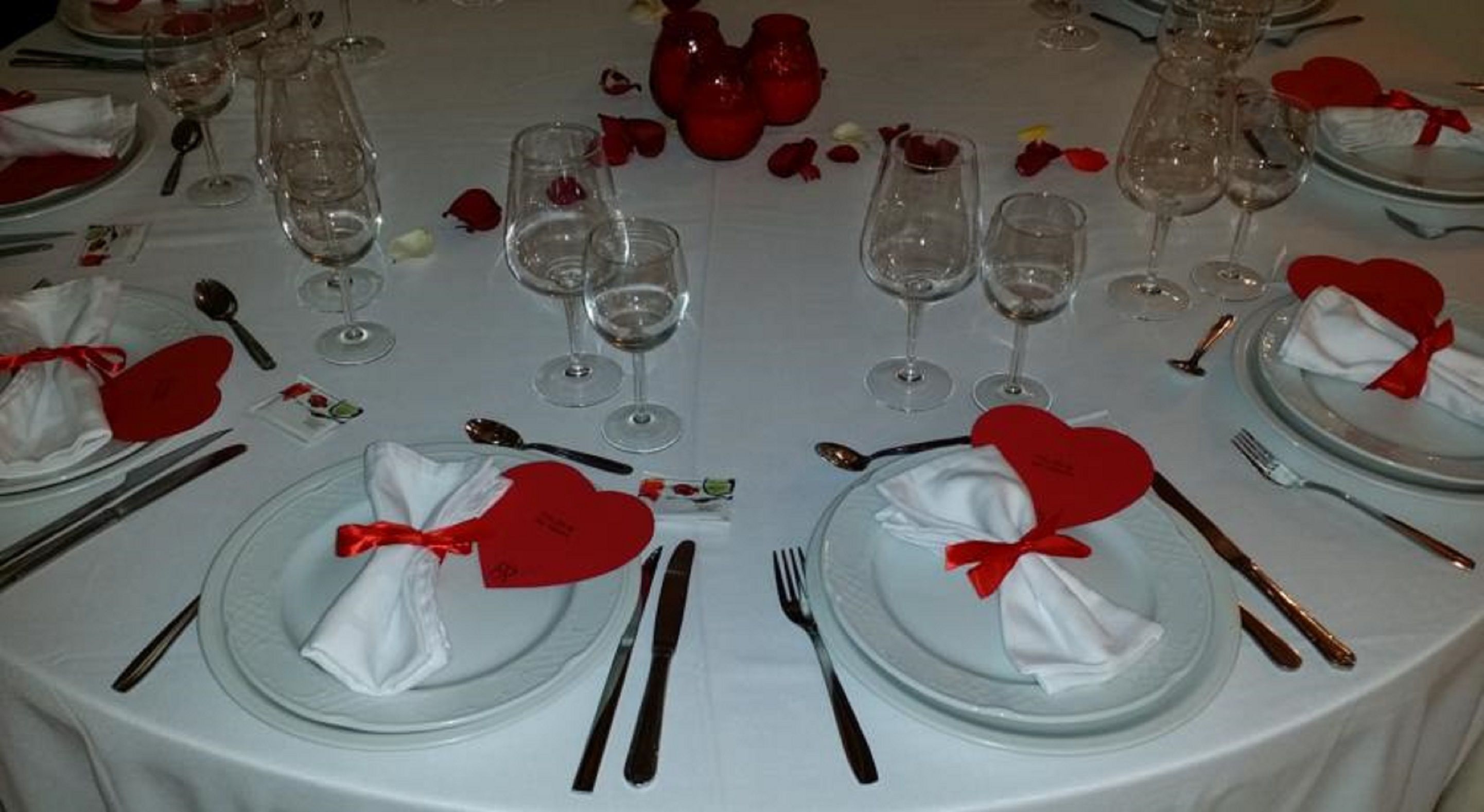 Una mesa redonda dispuesta para un evento formal, con manteles blancos, lazos de regalo en forma de corazón rojo y cristalería transparente.