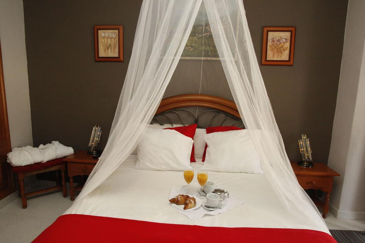 A neatly made bed with a red bedspread, white pillows, and a canopy. There are two bedside tables with candles and a framed picture on each. A breakfast tray with croissants, juice, and a cup is on the bed.