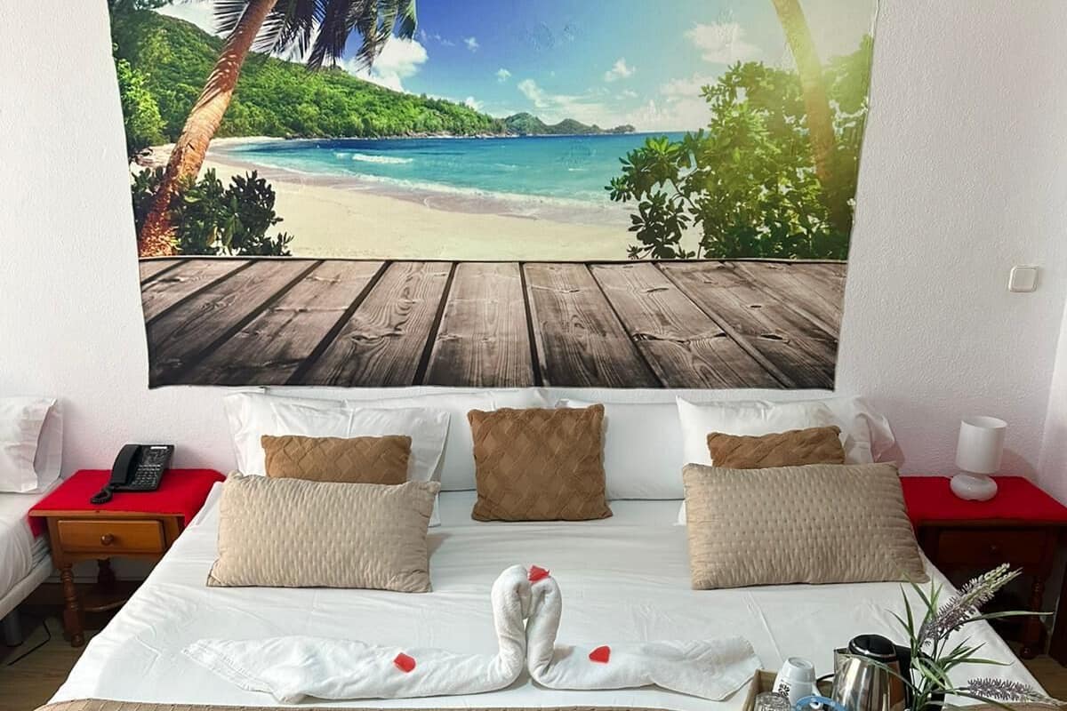 A bedroom with a large beach view mural, a neatly made bed with a towel and rose petals, and a bedside table with a lamp.