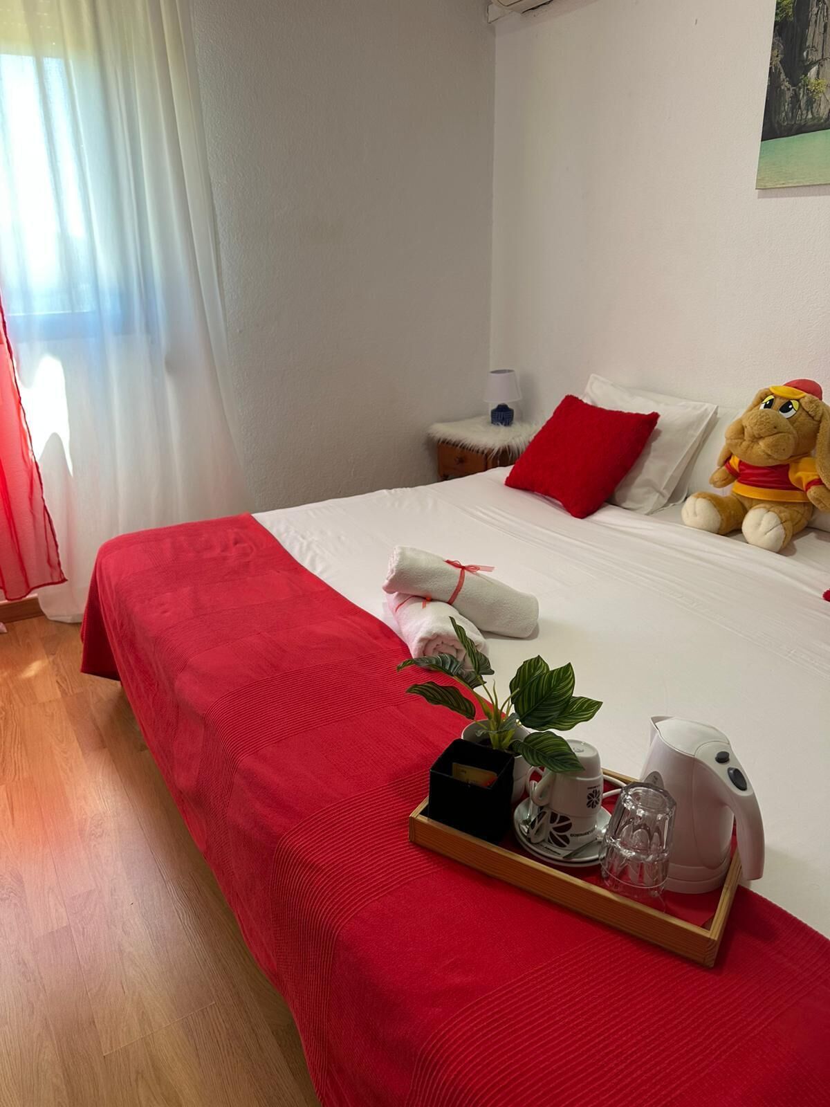 A hotel room with a red bedspread, a plush toy, a tray with a plant and a kettle, and a painting on the wall.