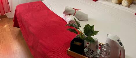 A hotel room with a red bedspread, a plush toy, a tray with a plant and a kettle, and a painting on the wall.