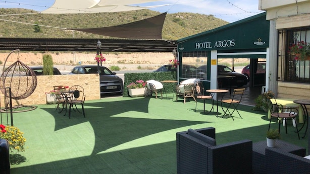 A hotel patio with outdoor seating, a car parked in front, and a desert landscape in the background. - Hotel Argos Murcia (Cehegin)