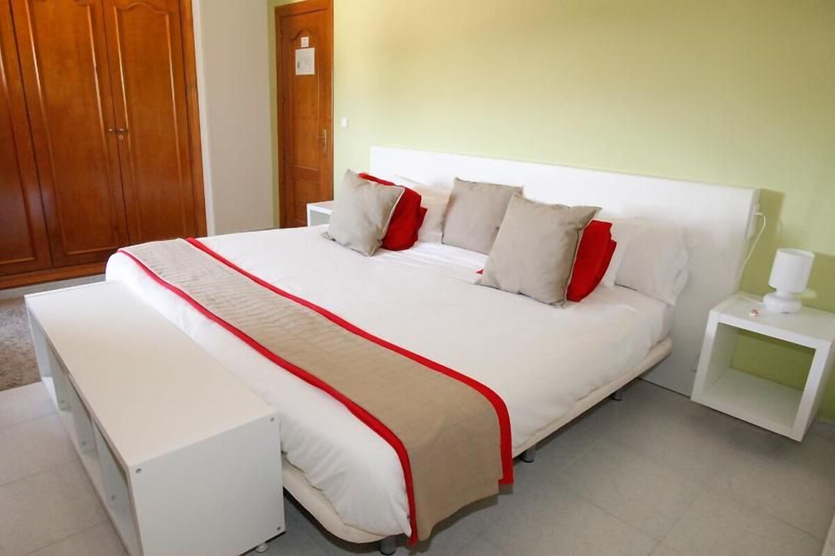 Una cama bien hecha con colcha roja y blanca, un armario de madera y una mesita de noche con lámpara.