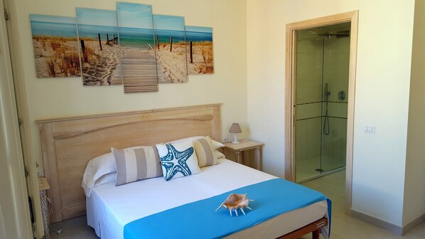 A bedroom with a wooden headboard, a bed with striped pillows, a blue blanket, a seashell decoration, a painting of a beach scene, and a glass-enclosed shower.