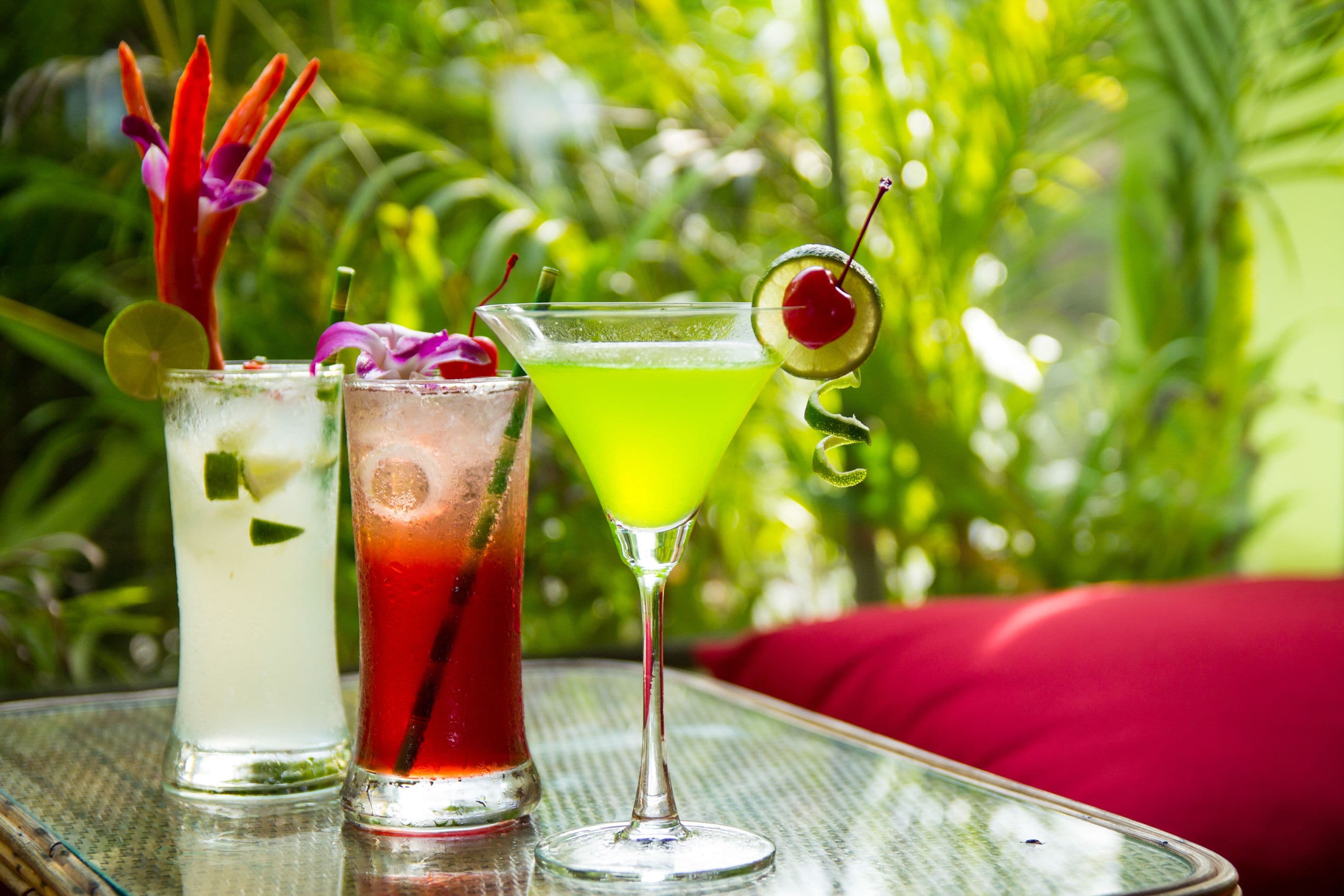 Three cocktails on a glass table with a red cushion in the background.