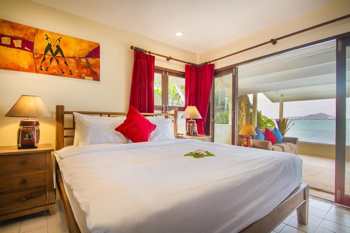 A hotel room with a large bed, red curtains, a painting on the wall, and a view of the sea.