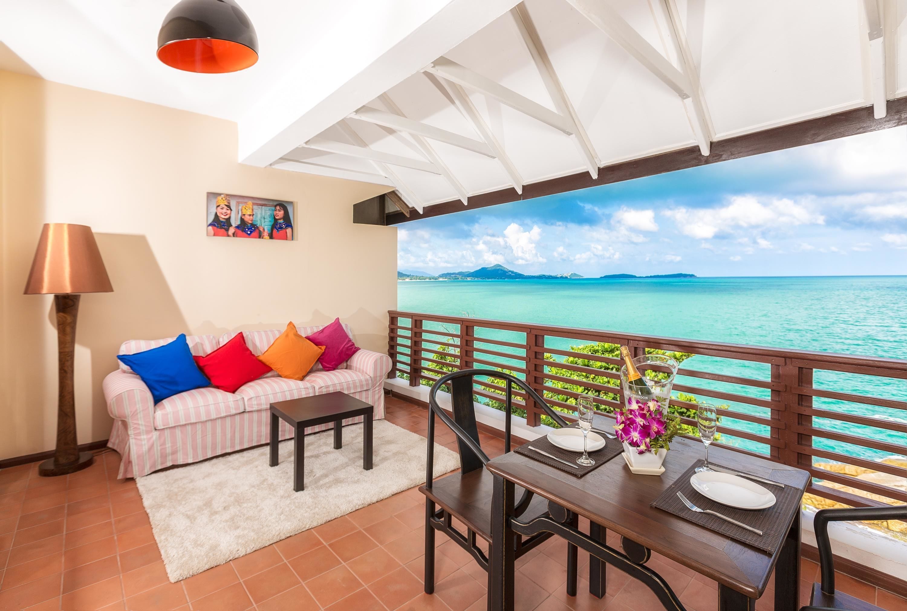 A balcony with a view of the ocean, a dining table set with a bottle and flowers, and a sofa with colorful cushions.