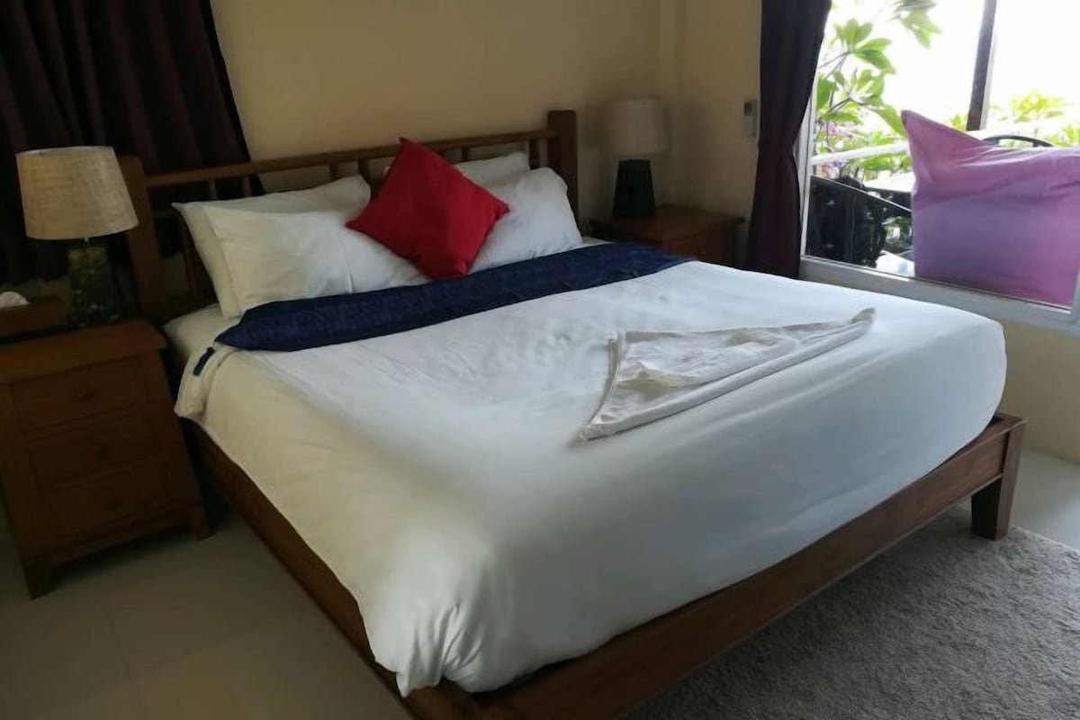 A neatly made bed with white bedding, a blue throw, and a red pillow. There is a wooden nightstand with a lamp and a small table beside the bed.