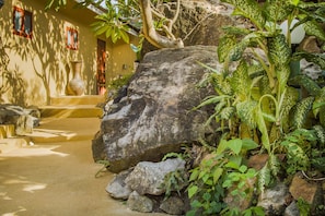 Uma trilha de pedra leva a um edifício, com uma grande rocha em primeiro plano e vegetação exuberante ao redor.