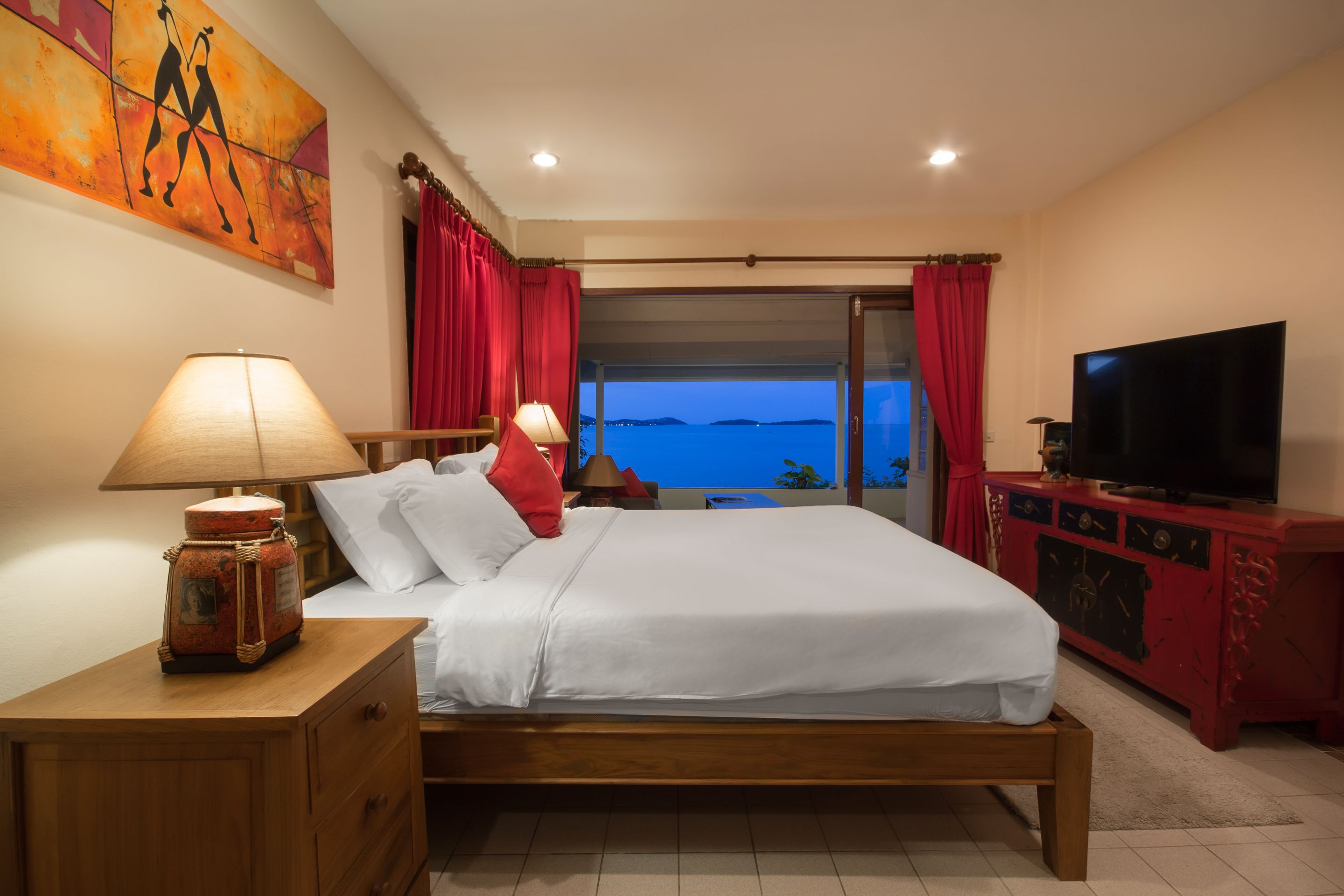 Quarto com cama, televisão, mesa de cabeceira, abajur e vista para o mar.