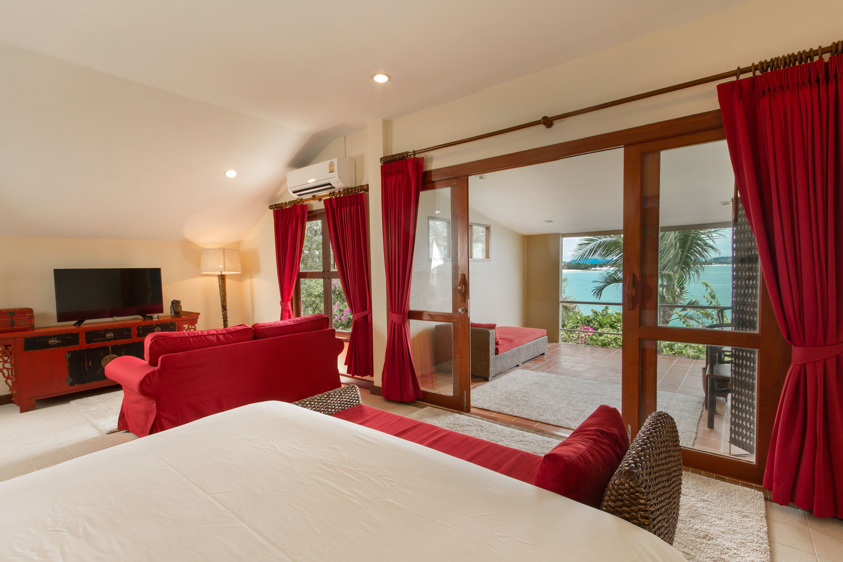 A hotel room with a large bed, red curtains, a TV, and a balcony with a view of the sea.