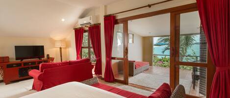 A hotel room with a large bed, red curtains, a TV, and a balcony with a view of the sea.