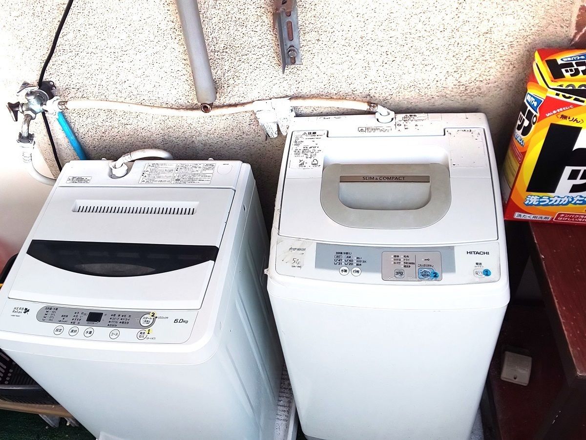 Two white washing machines with control panels, a metal pipe, and a cardboard box with Japanese text.