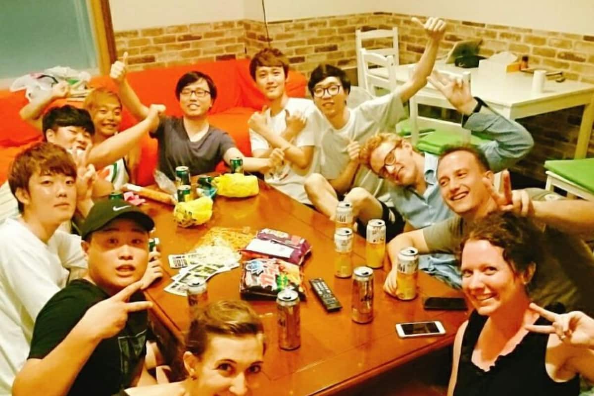 Eine Gruppe von Menschen sitzt an einem Tisch mit Snacks und Getränken in einem Raum mit Backsteinwänden und einem grünen Sofa.