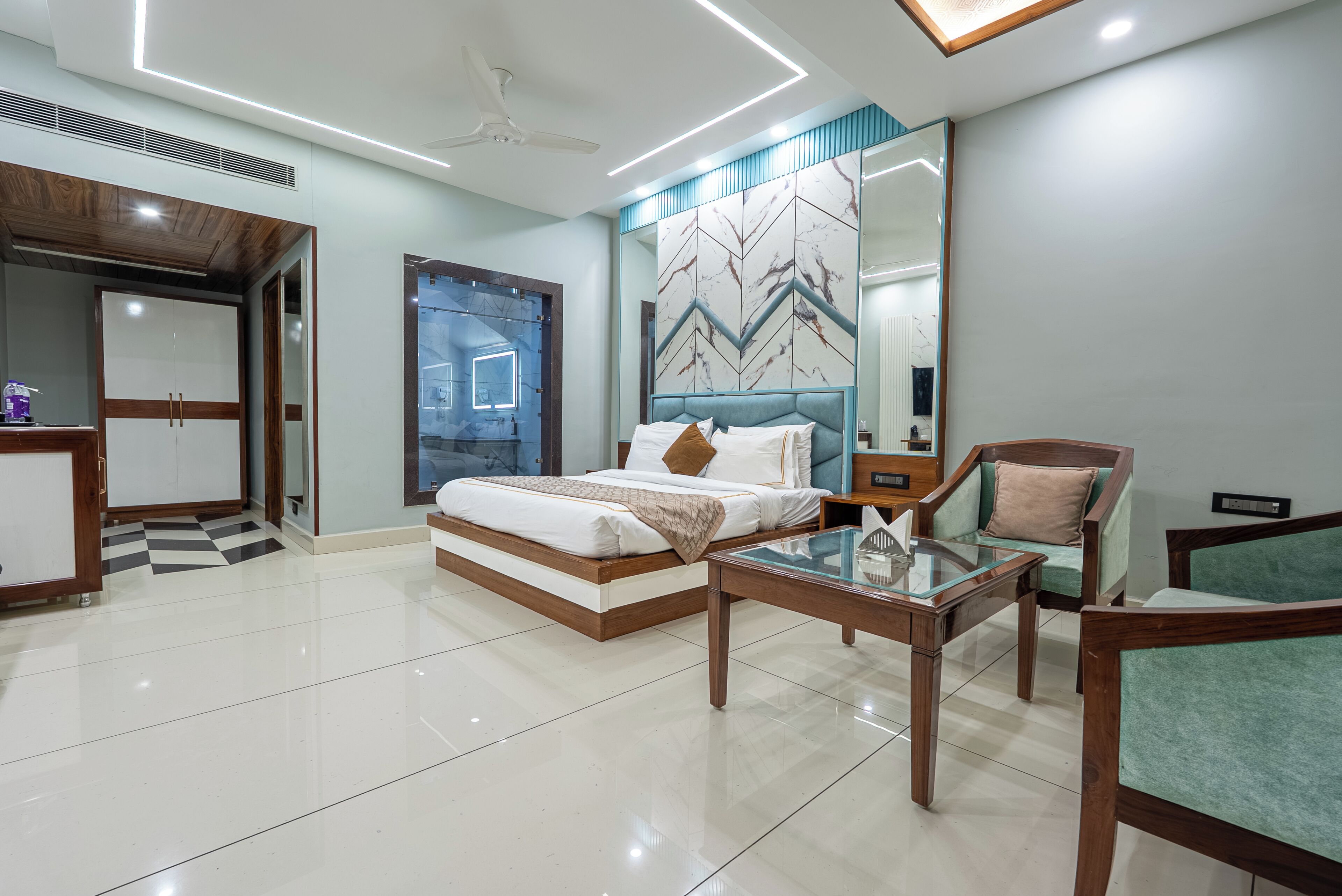 A modern hotel room with a bed, a glass coffee table, two armchairs, a large mirror, and a ceiling fan.