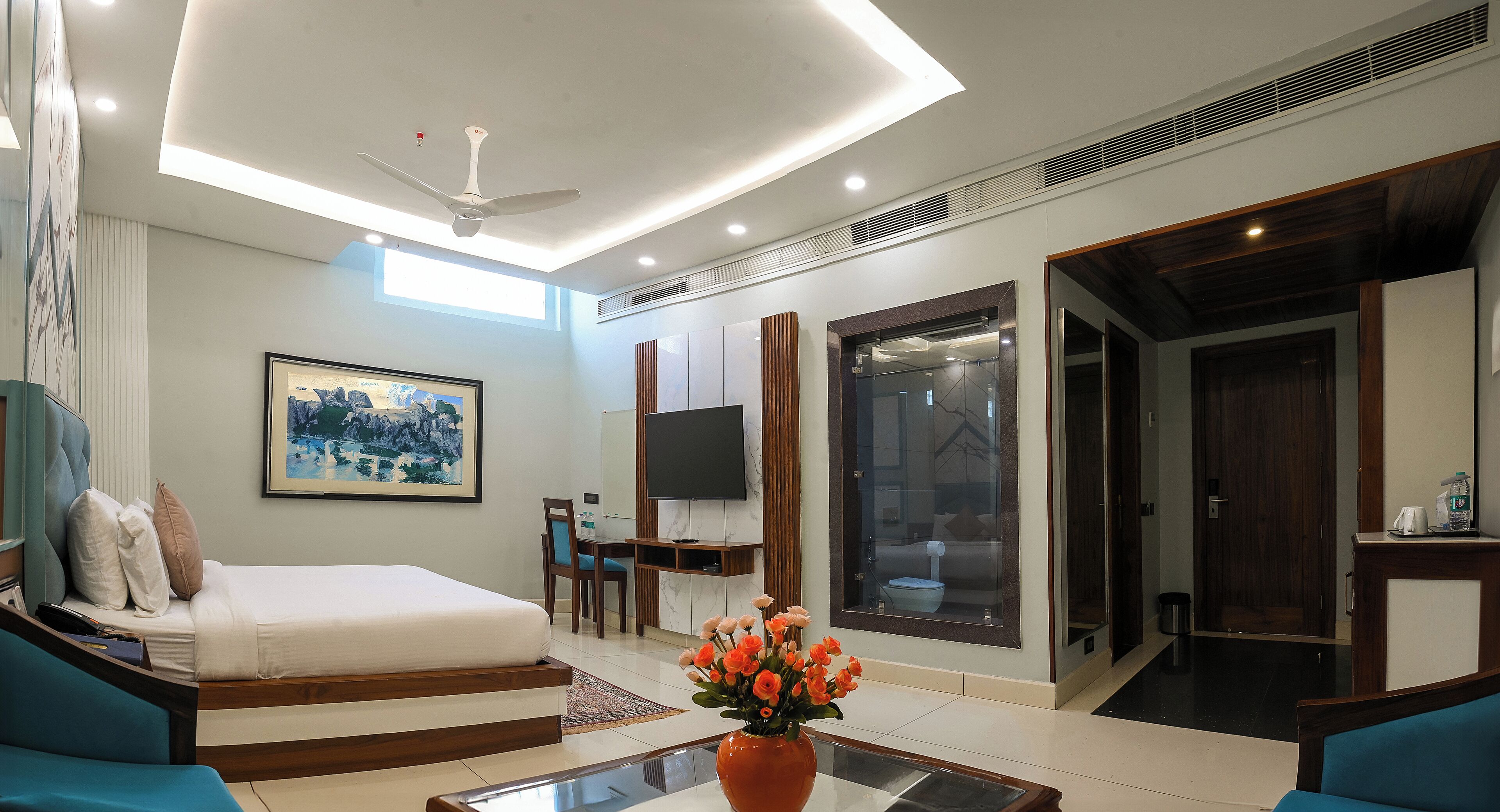 Habitación de hotel moderna con cama, sofá, televisión y baño con puerta de vidrio.