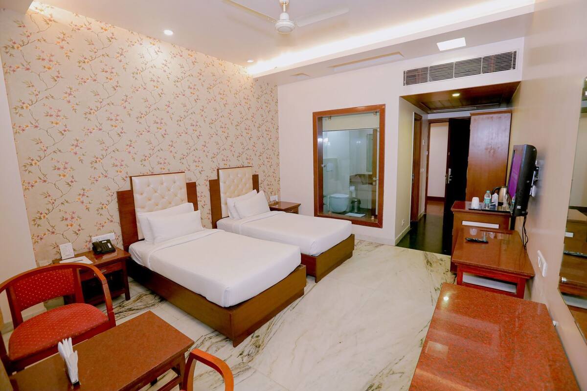 A hotel room with two beds, a desk, a chair, a TV, and a bathroom visible through a glass door.