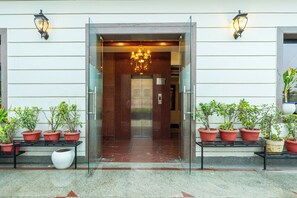 A hotel entrance with a glass door, a chandelier, potted plants, and a bench. - Leela Grande Hotel (Karnal)
