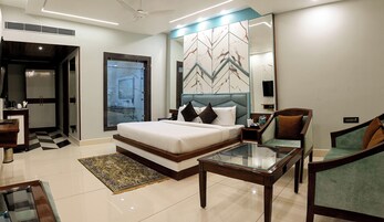 Una habitación de hotel moderna con una cama grande, una mesa de centro de cristal, una zona de estar con una silla y un baño visible a través de una puerta abierta.