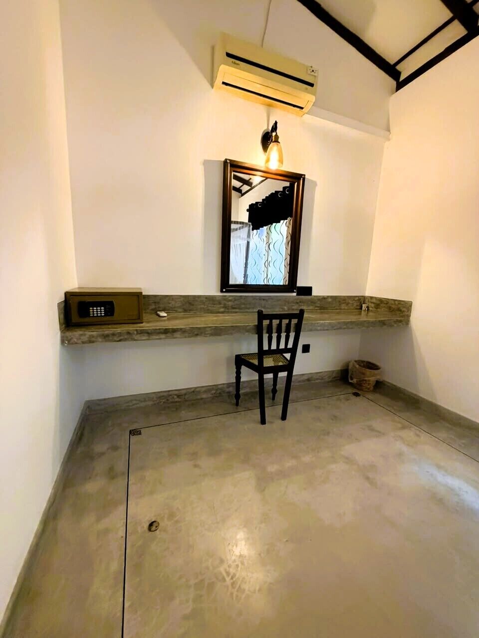 A room with a concrete floor, a chair, a small table with a device, a wall-mounted air conditioner, and a framed picture with a light.