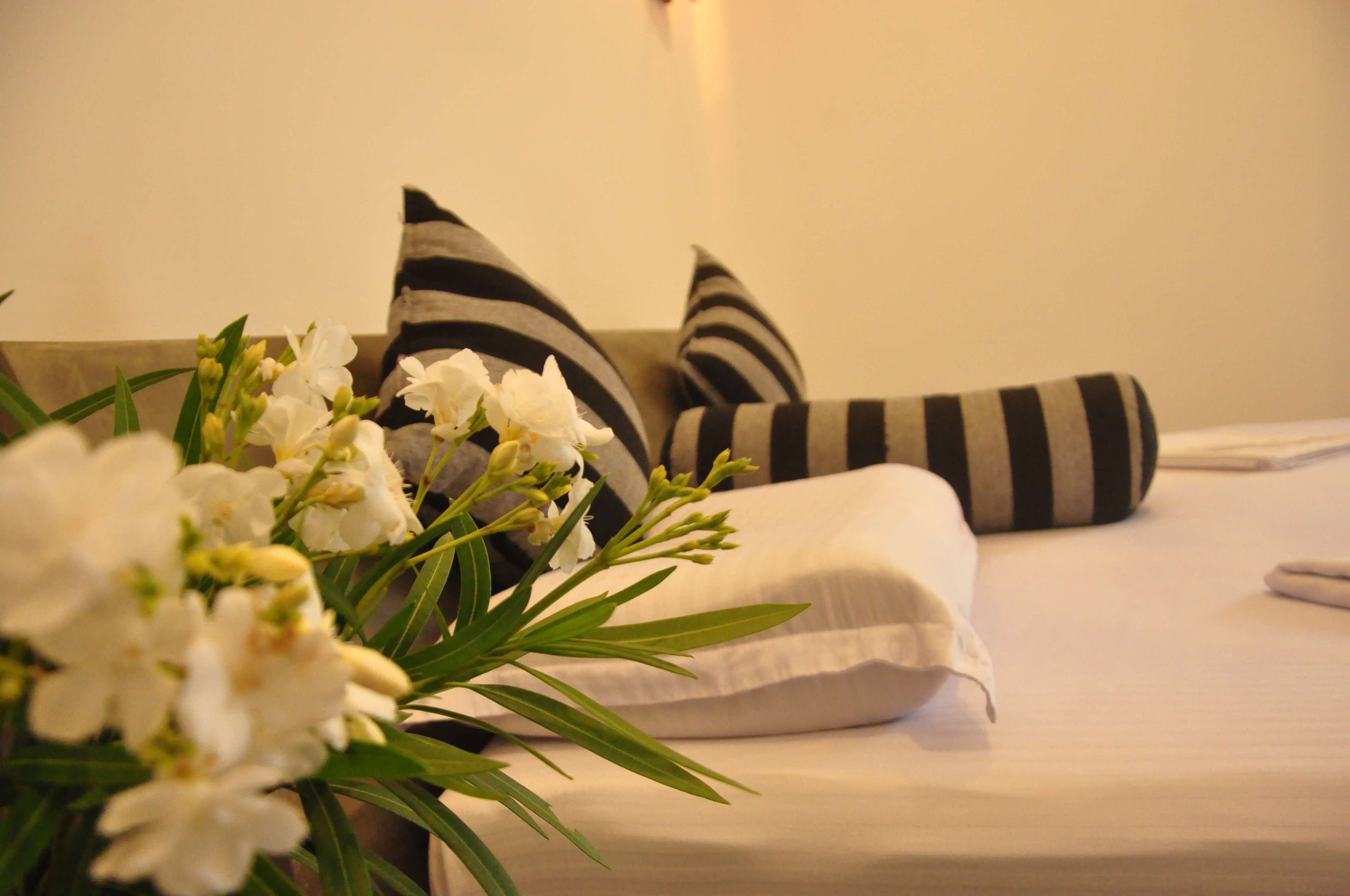 A neatly made bed with white linens, striped pillows, and a bouquet of white flowers.