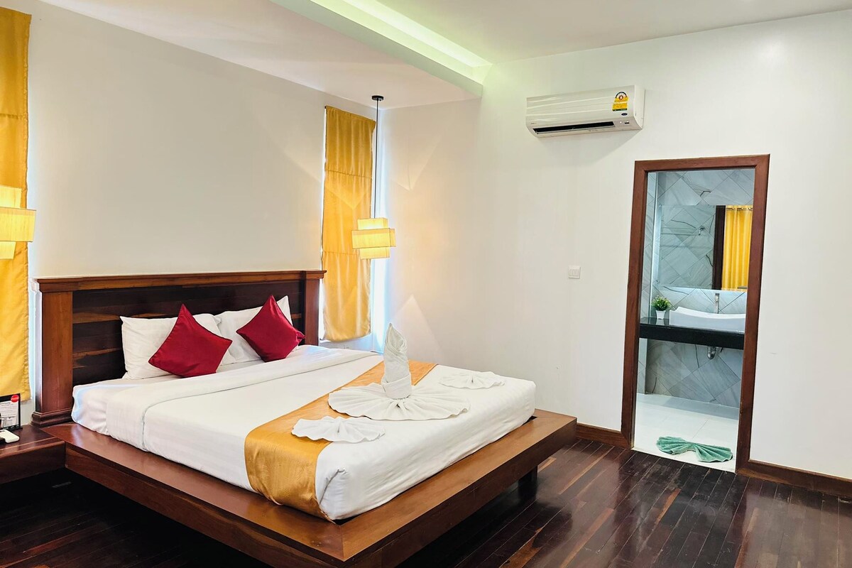 Habitación de hotel con cama de madera, ropa de cama blanca, almohadas rojas y blancas, una lámpara de noche, equipo de aire acondicionado y un baño con espejo y lavamanos.