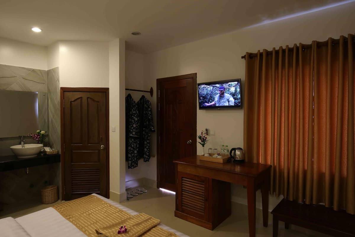 A hotel room with a bed, a TV, a wooden desk, a bathroom with a sink and mirror, and a door leading to another room.
