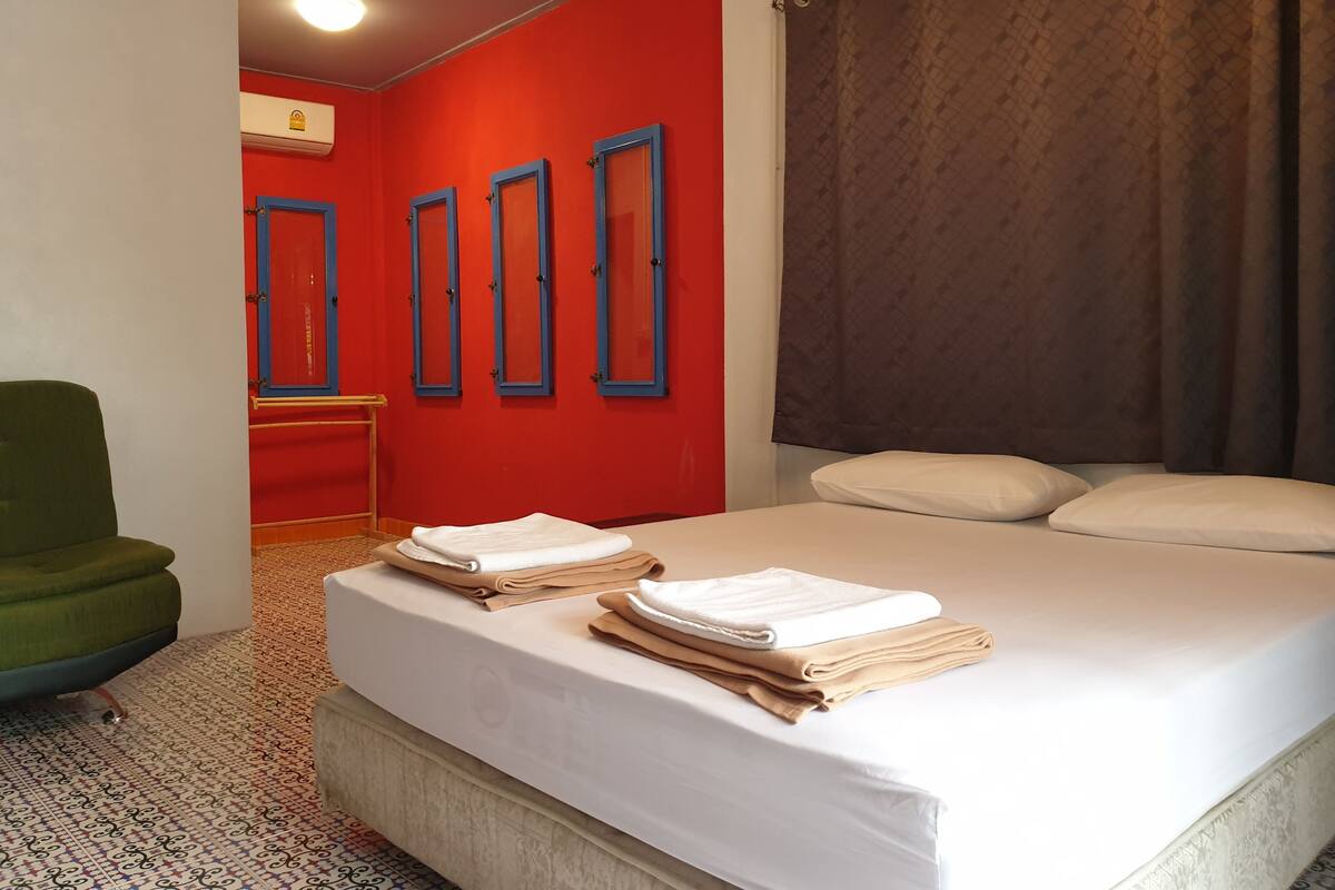 A hotel room with a bed, a green chair, a red wall, and a brown curtain.