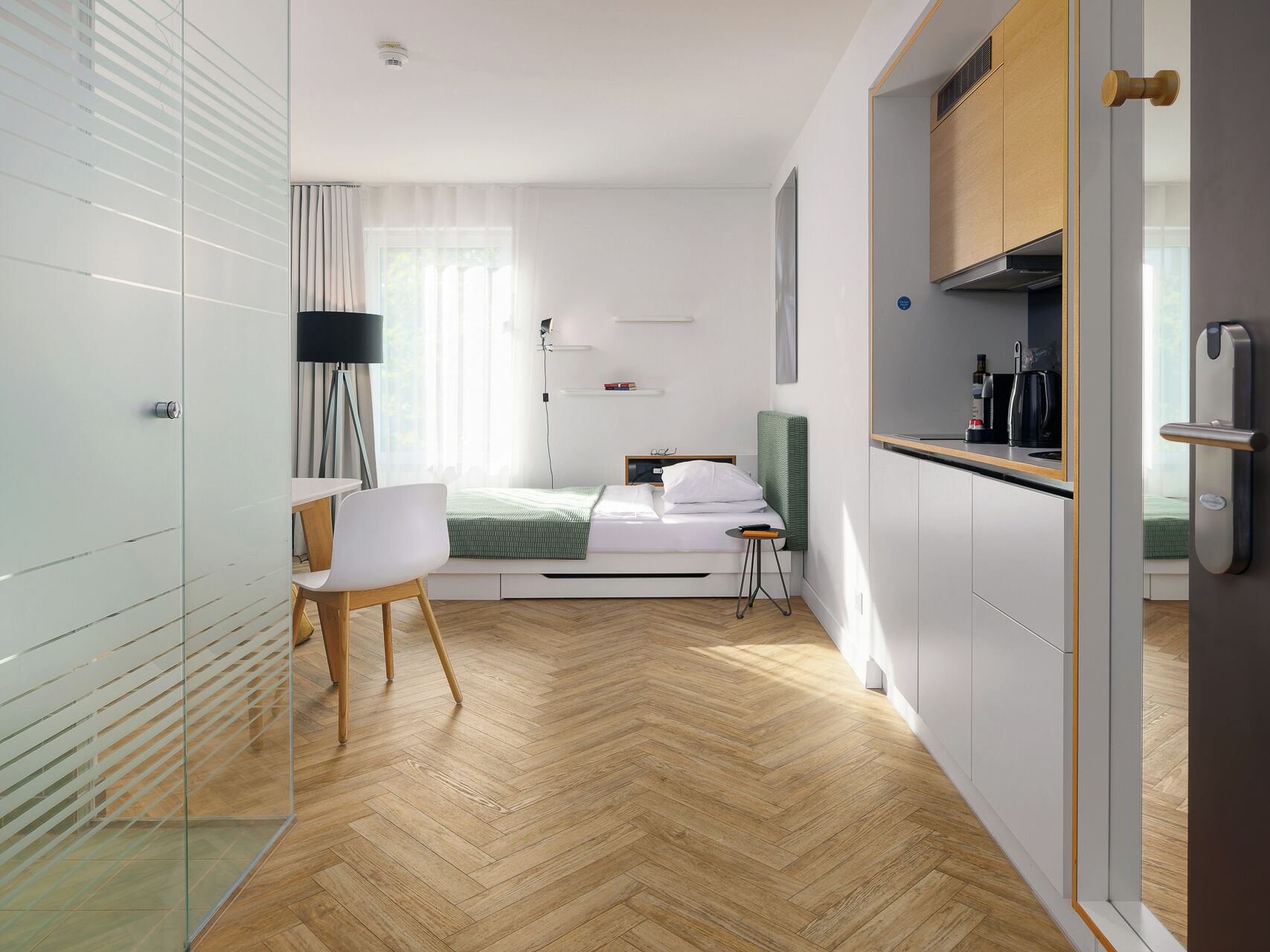 Habitación de hotel moderna con cama, mesita de noche, silla y una pequeña cocina.