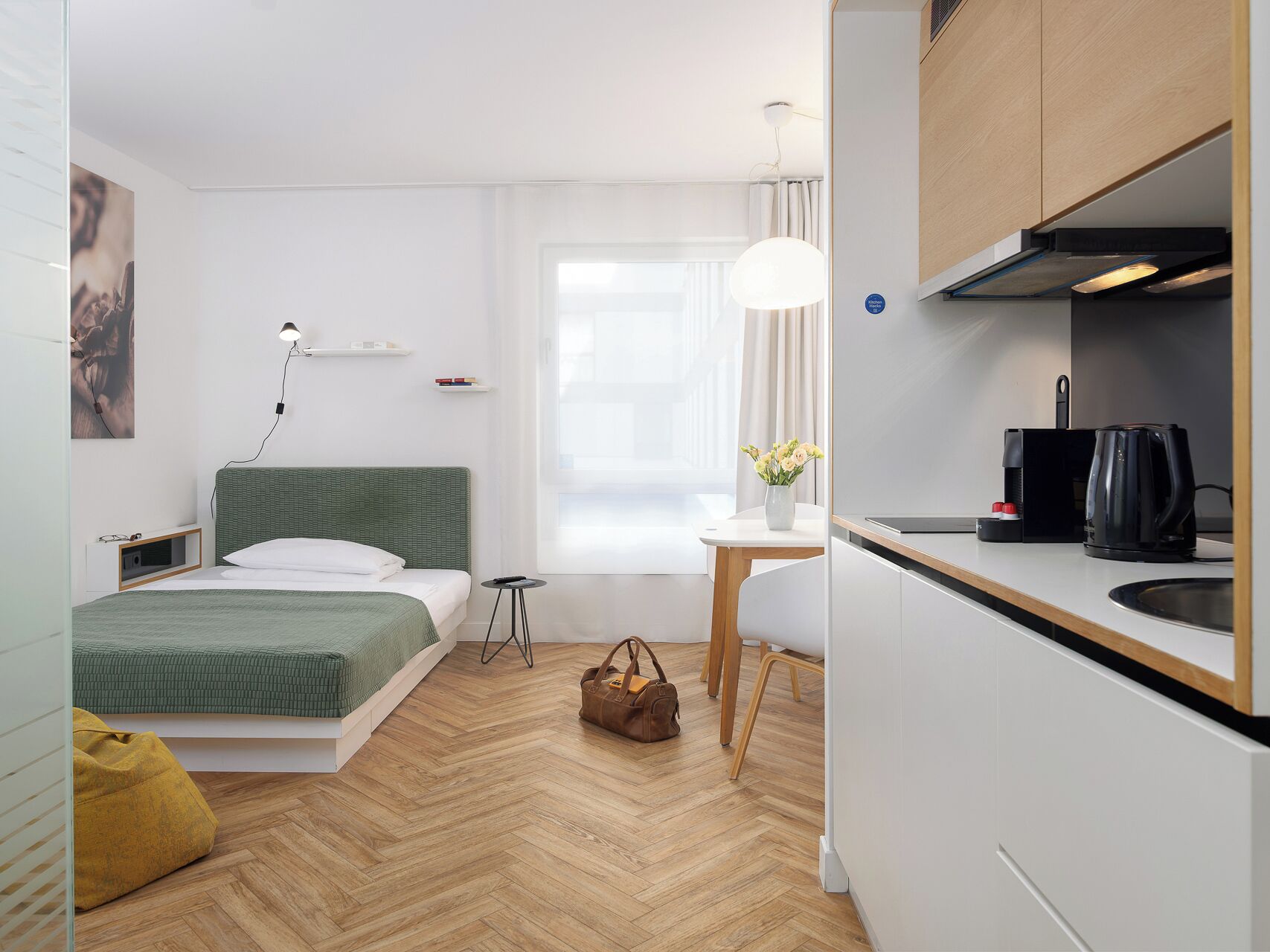 A modern hotel room with a bed, bedside table, lamp, and a kitchenette with a coffee maker.