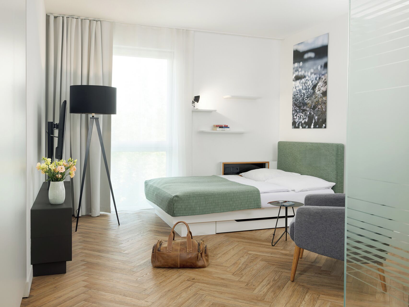 A modern hotel room with a bed, a desk, a chair, a lamp, and a painting on the wall.