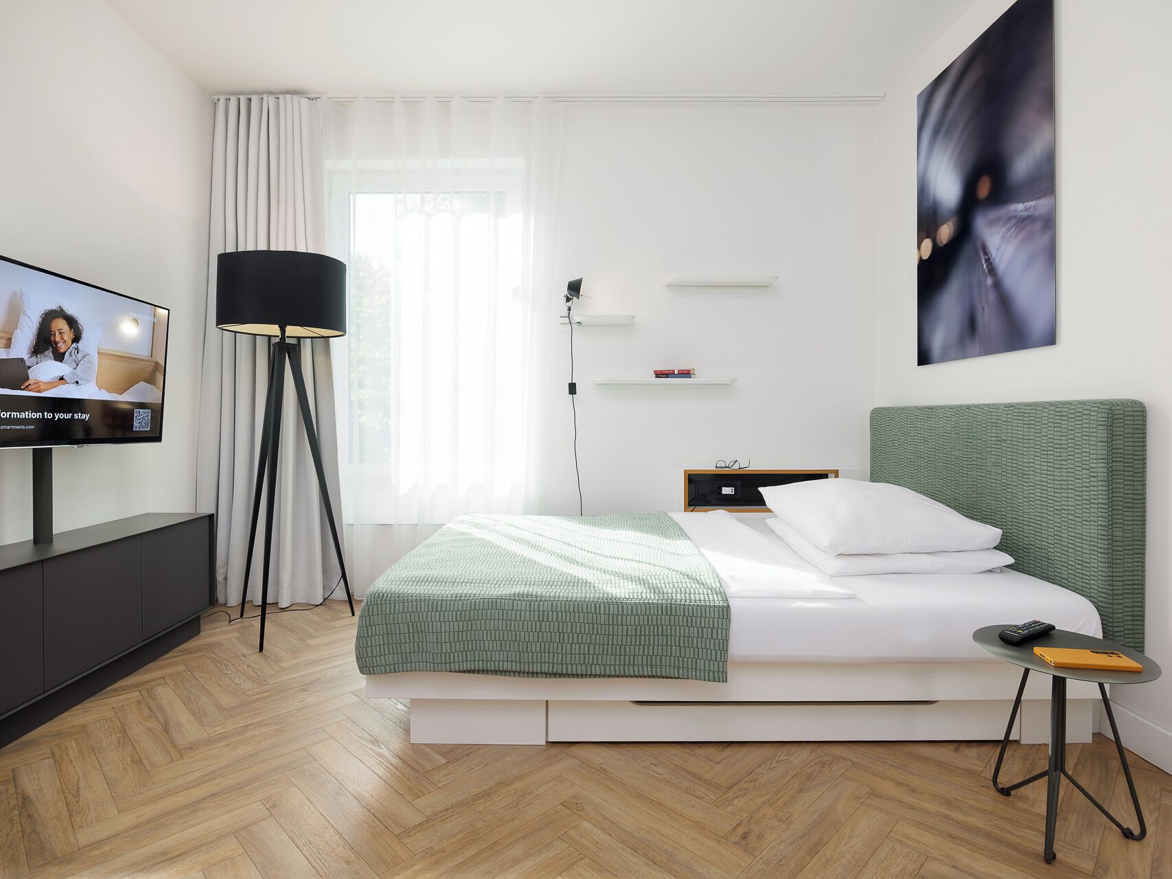 A modern hotel room with a bed, bedside table, lamp, TV, and a view of the outdoors.