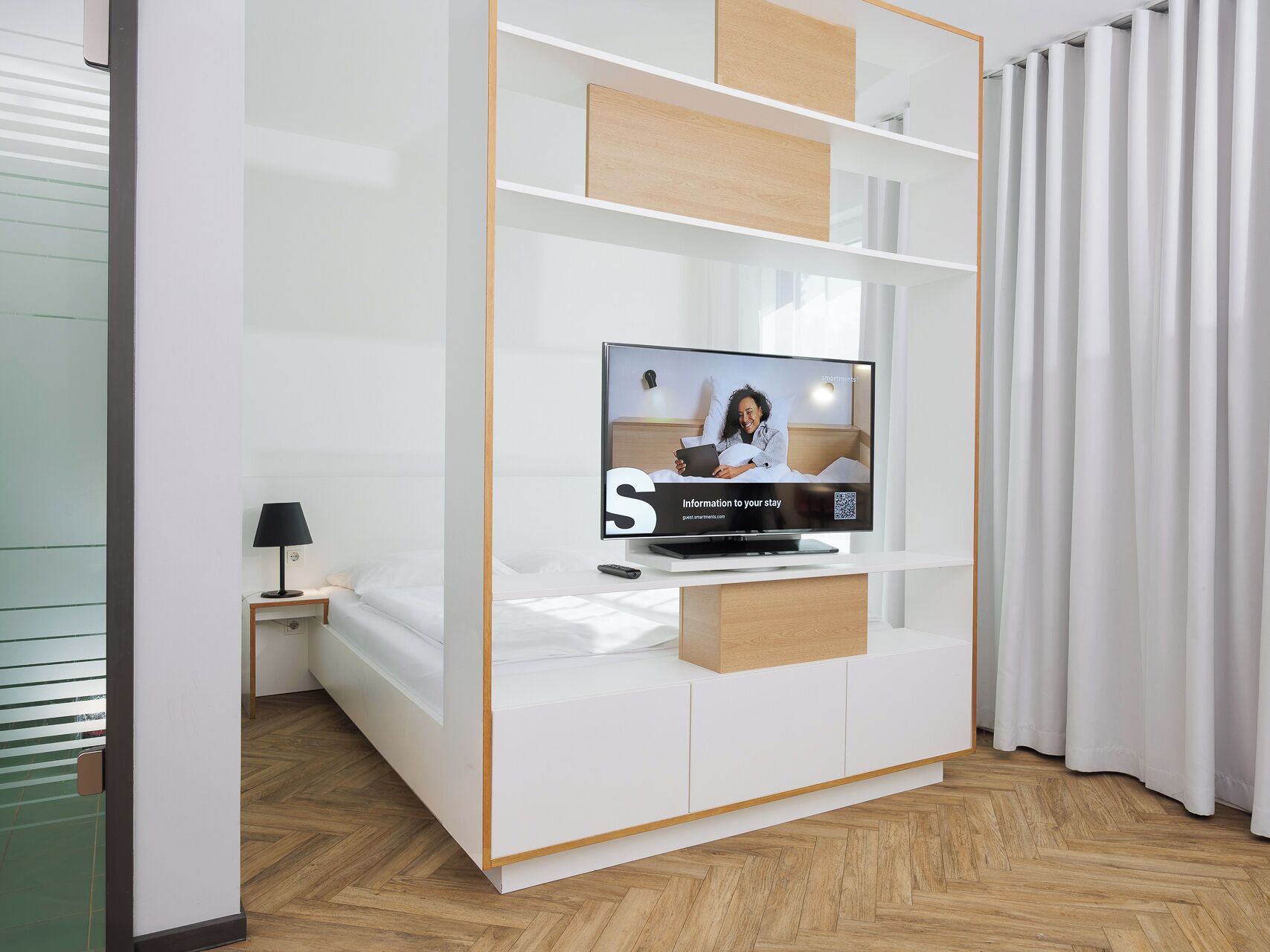 Ein modernes Hotelzimmer mit einem Flachbildfernseher auf einem Holzboden, einem Nachttisch mit Lampe und einem Bett mit weißer Bettwäsche.