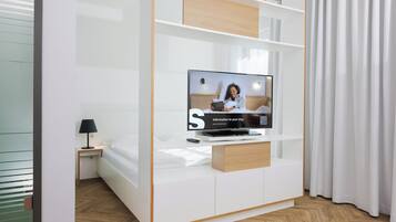 Ein modernes Hotelzimmer mit einem Flachbildfernseher auf einem Holzboden, einem Nachttisch mit Lampe und einem Bett mit weißer Bettwäsche.