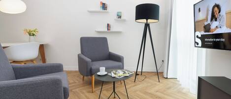 Un salon moderne avec un canapé gris, une table basse sur laquelle sont posés des magazines, un lampadaire et un téléviseur qui affiche un message de bienvenue.