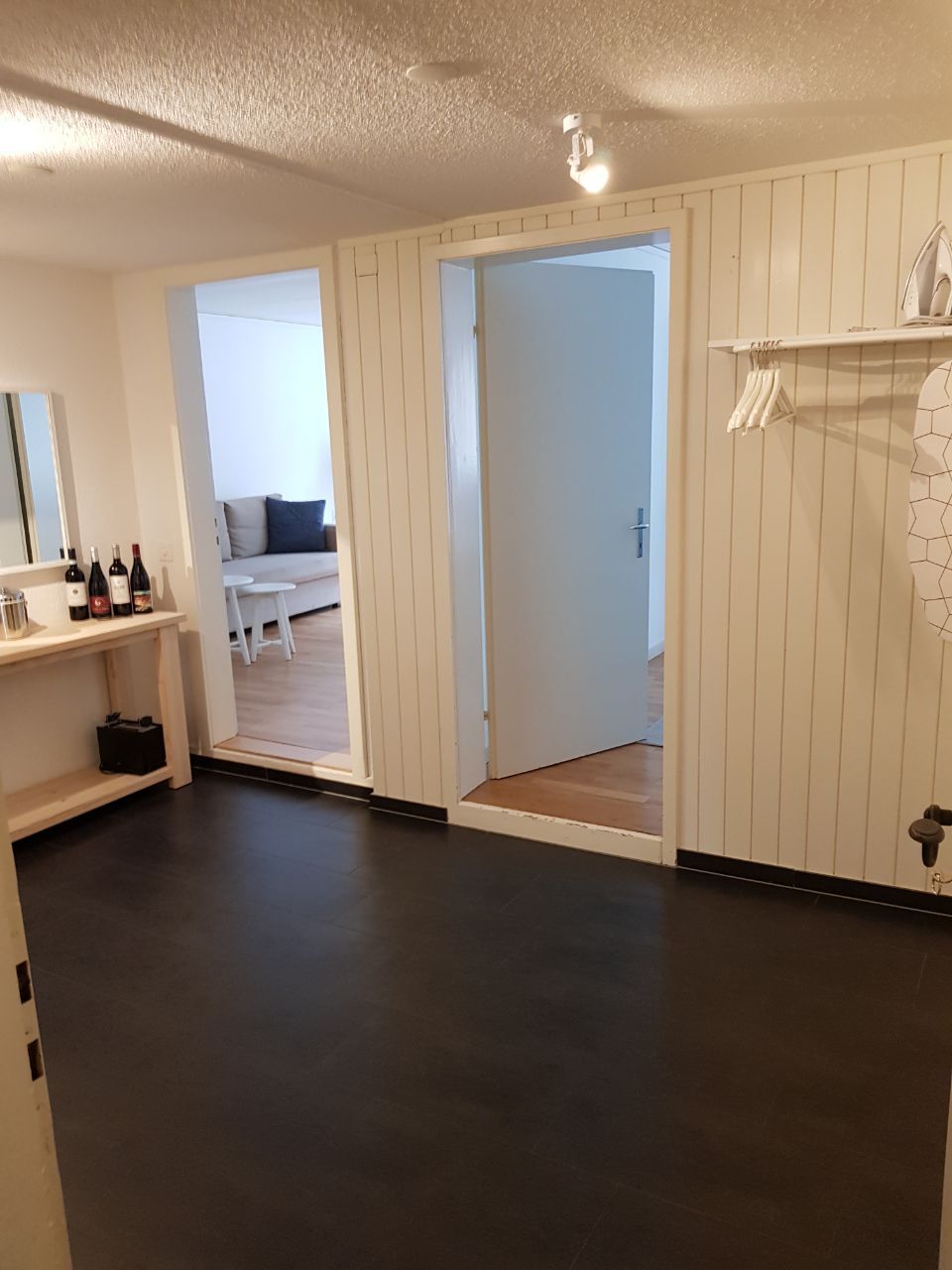 A room with a dark floor, a white door, a white wall with a shelf, and a white ceiling.