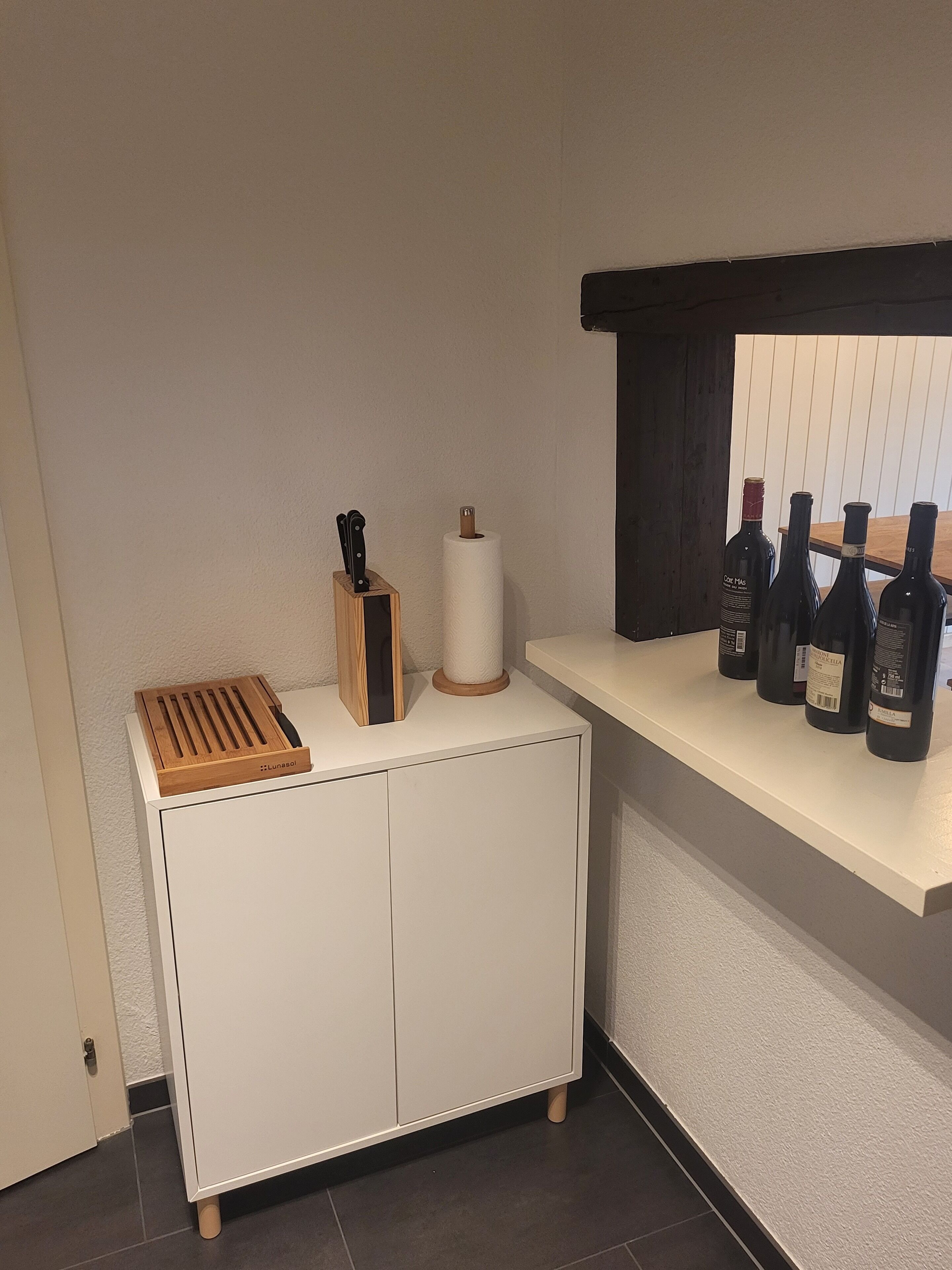 A kitchen with a white cabinet, a wooden cutting board, a knife block, a roll of paper towels, and a shelf with wine bottles.