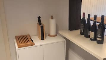 A kitchen with a white cabinet, a wooden cutting board, a knife block, a roll of paper towels, and a shelf with wine bottles.