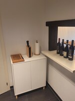 Una cocina con un armario blanco, tabla de cortar de madera, bloque de cuchillos, rollo de servilletas de papel y una estantería con botellas de vino.