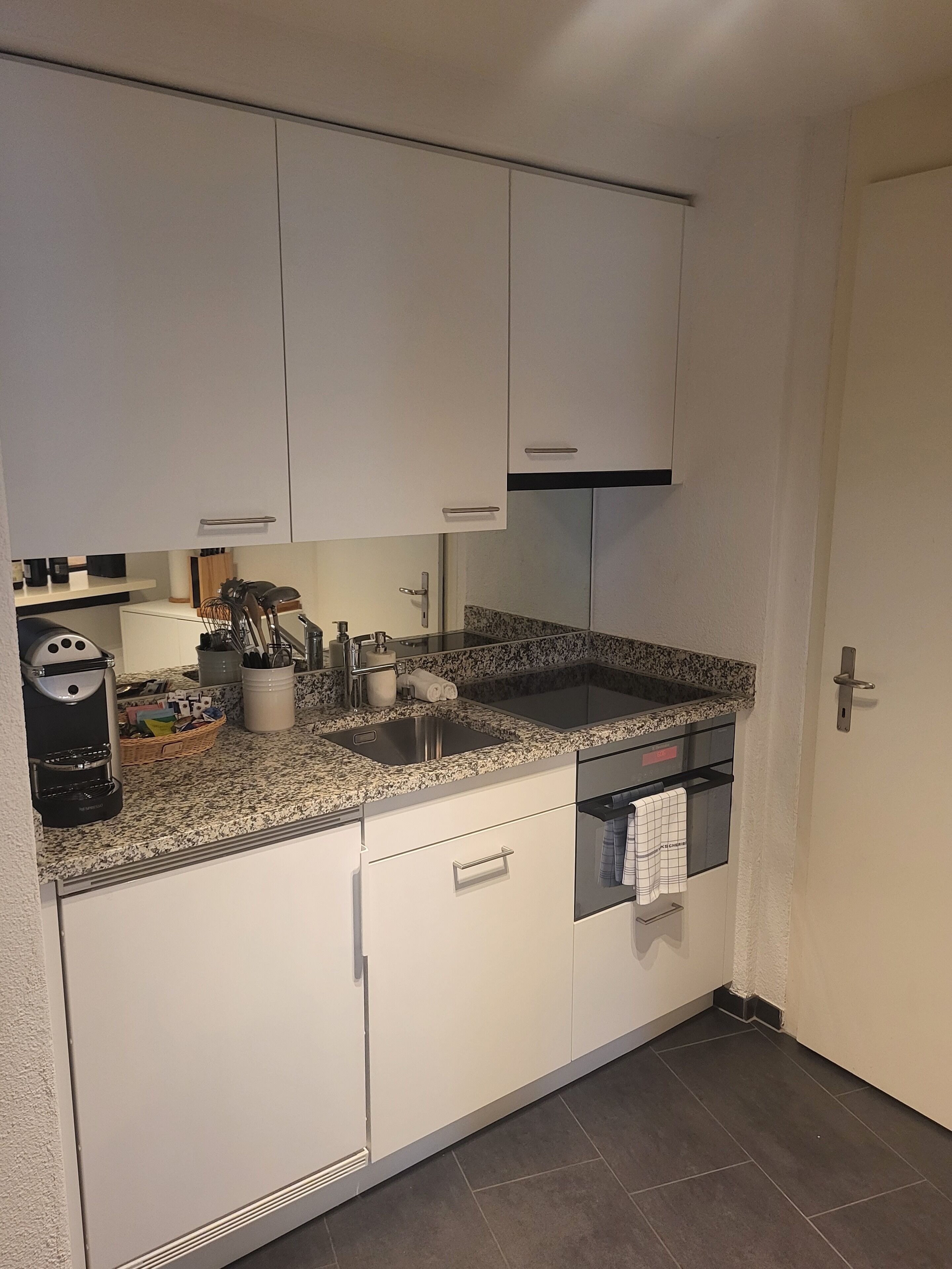 Une cuisine compacte avec des meubles blancs, un plan de travail en granit, un four encastré et une machine à café.