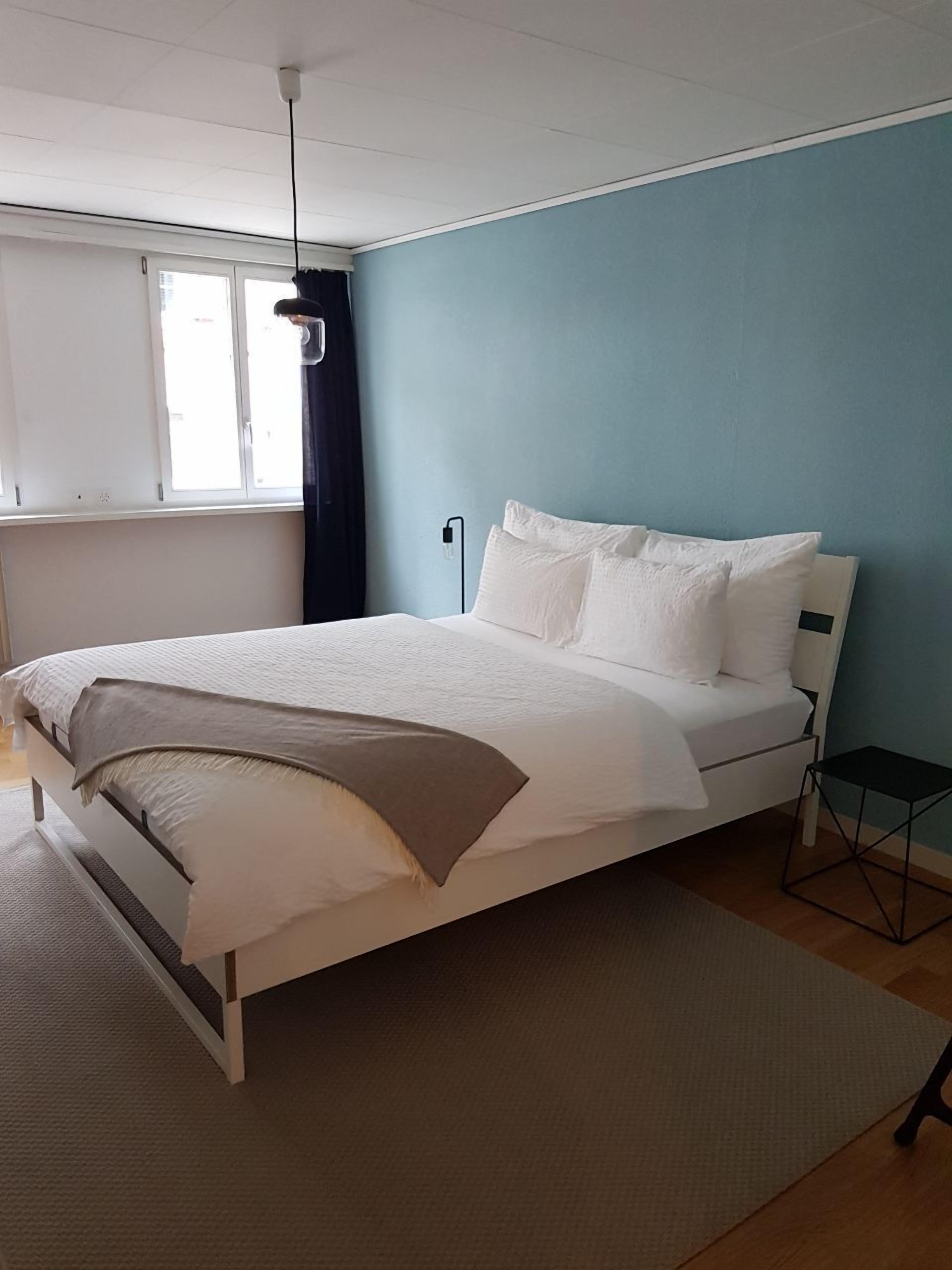 A bedroom with a bed, a bedside table, a window with curtains, and a wall-mounted light fixture.