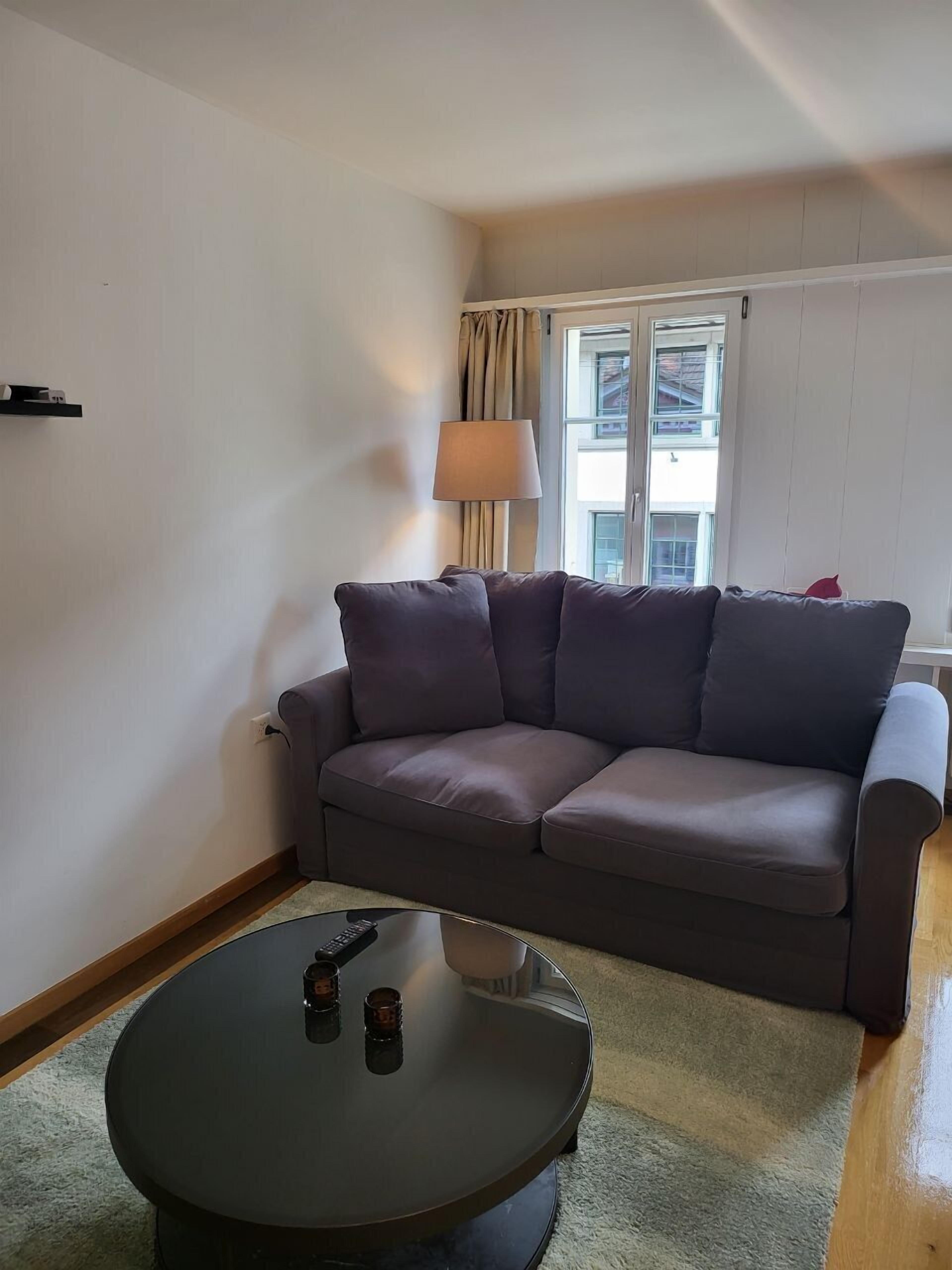 Una sala de estar con un sofá, una mesa redonda de centro, una lámpara y una ventana con cortinas.