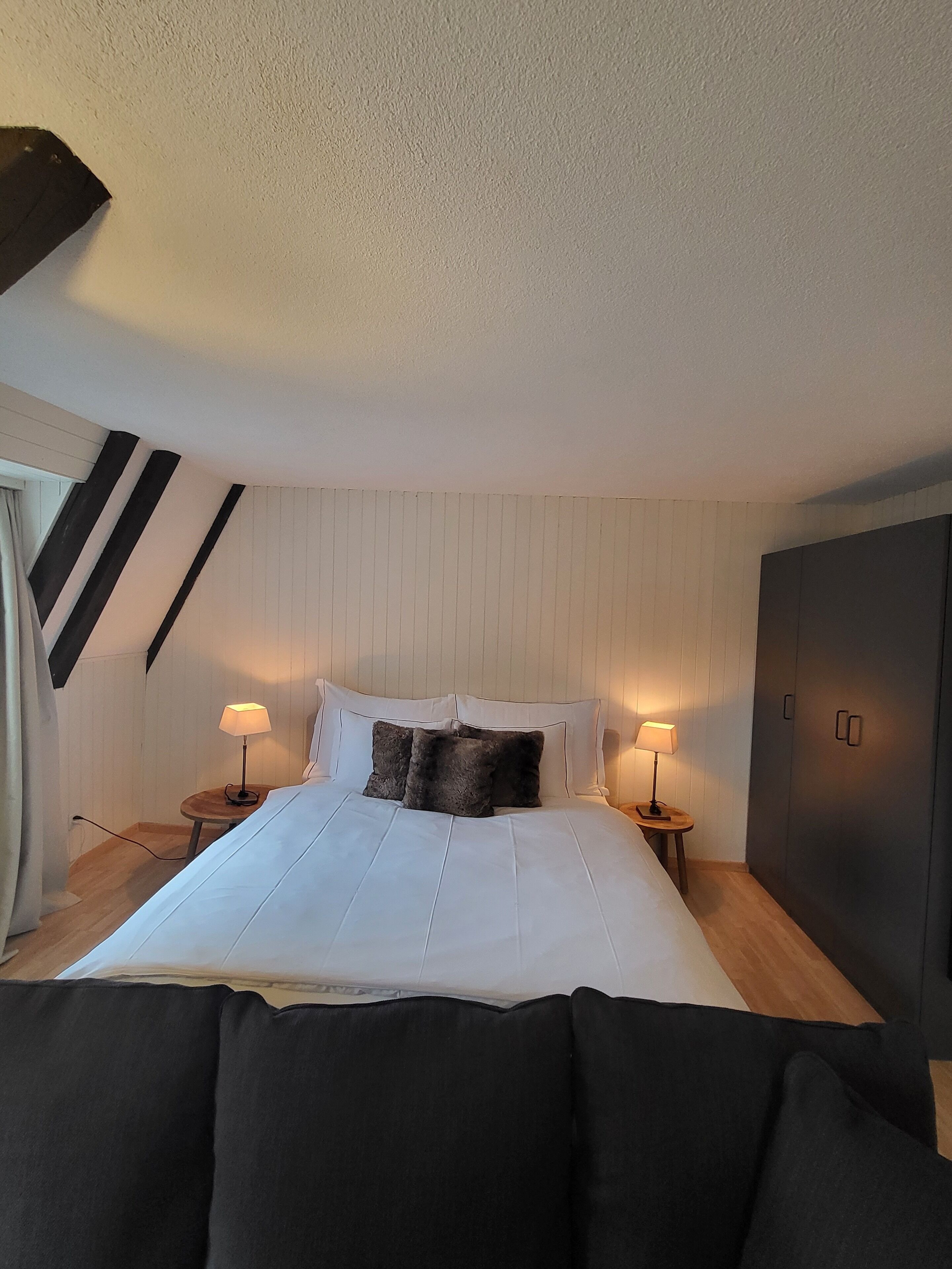 A bedroom with a large bed, two bedside tables with lamps, a wardrobe, and a sloped ceiling.