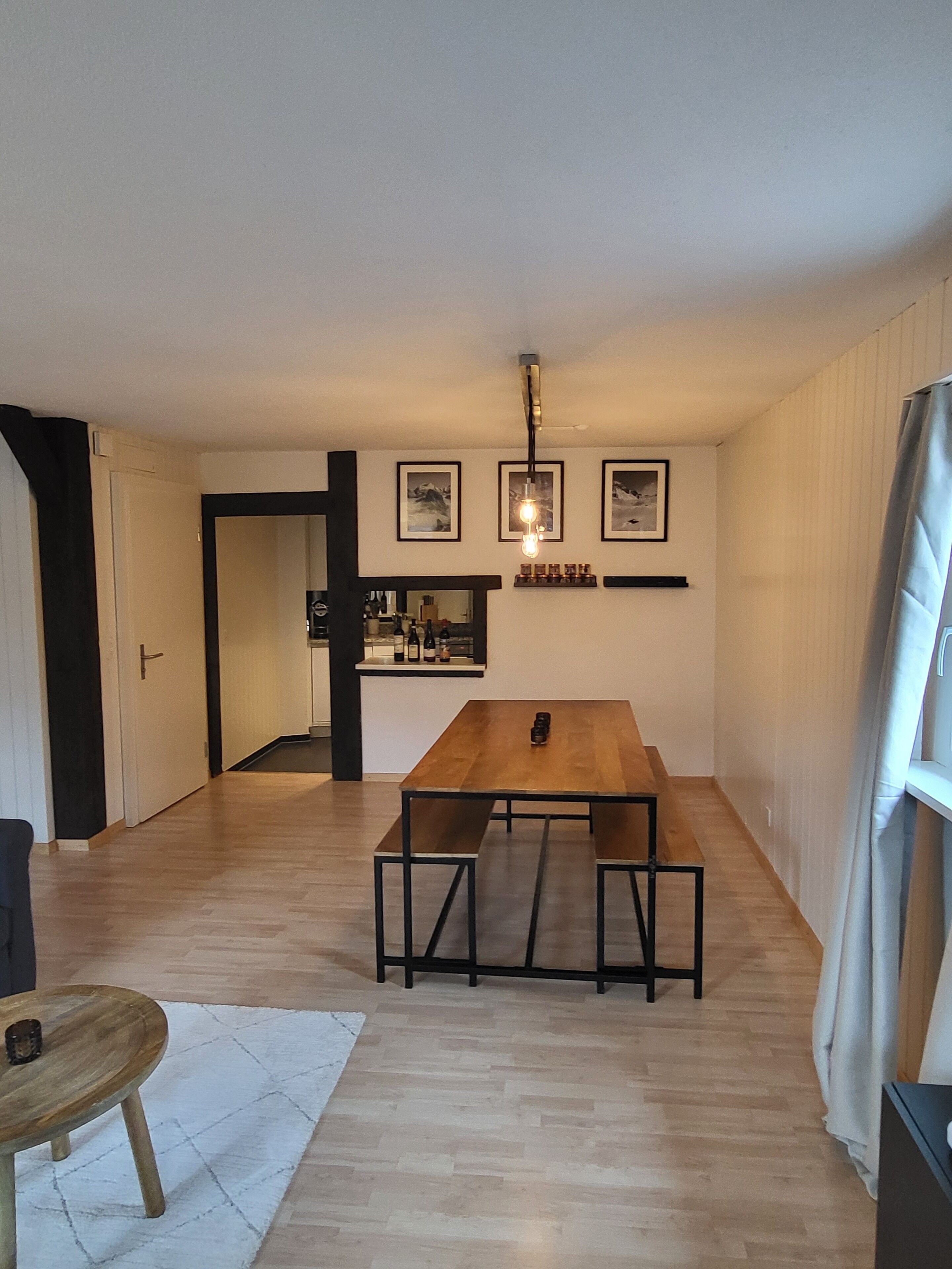 A dining area with a wooden table and benches, a kitchen in the background, and framed pictures on the wall.