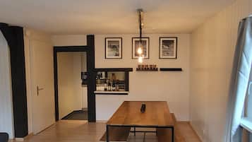 A dining area with a wooden table and benches, a kitchen in the background, and framed pictures on the wall.
